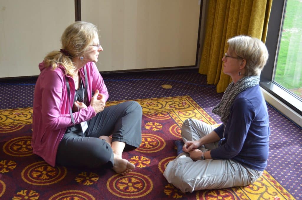 Two women sitting and talking on patterned carpet.